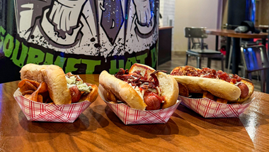 Three loaded hot dogs on wooden table with restaurant in the background.